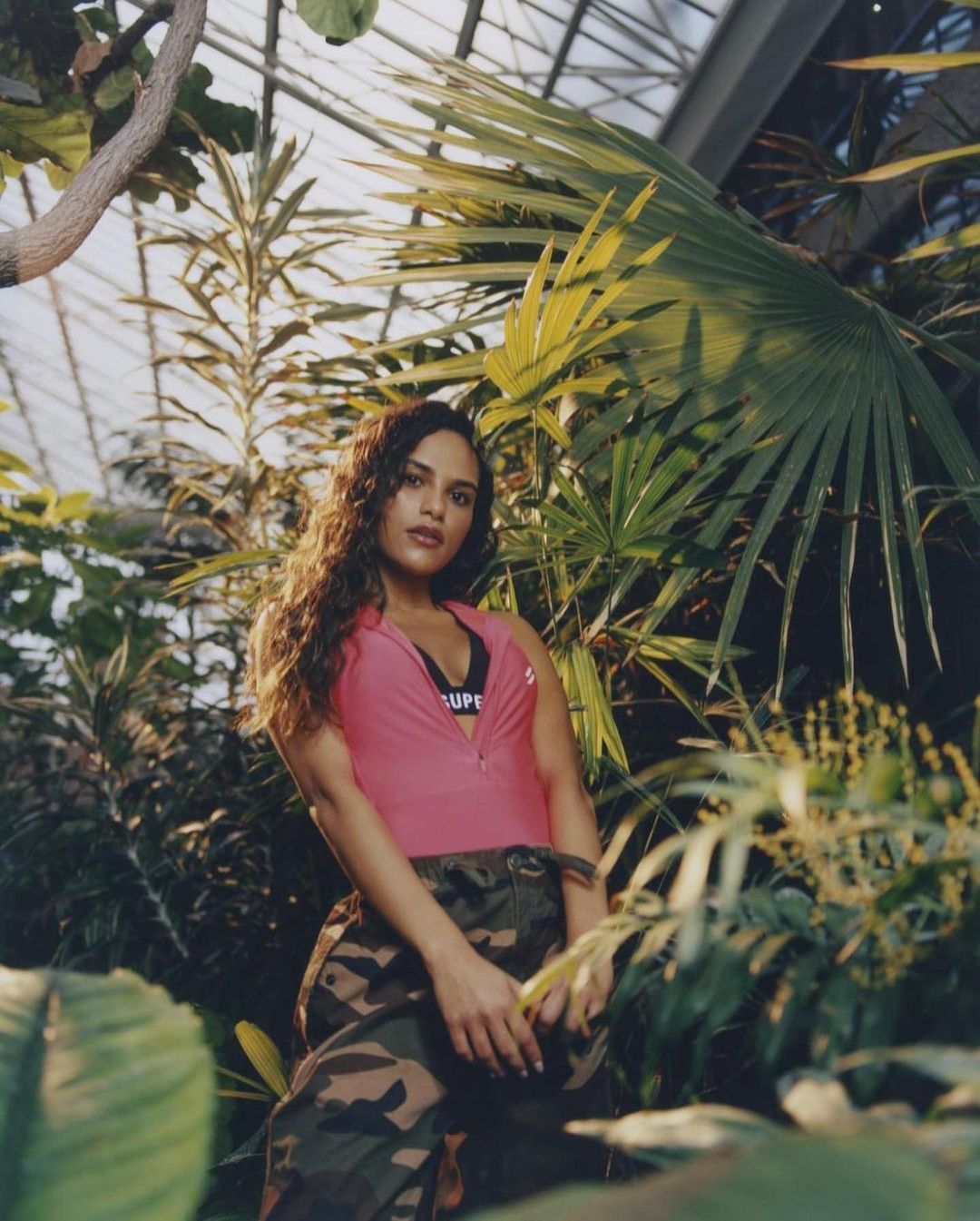 Woman with long, curly hair poses in a pink top and camo pants amidst lush greenery in a greenhouse-like environment with natural light.