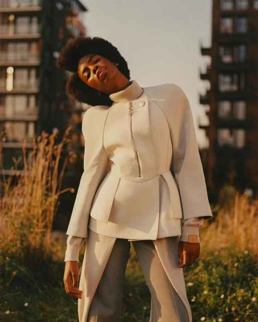 Fashionable woman with afro hairstyle poses outdoors wearing a cream-colored jacket and pants on a sunny day in an urban setting.