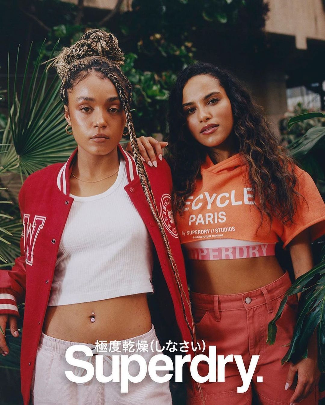 Two women pose in front of greenery, wearing Superdry branded clothing. One wears a red jacket and the other an orange cropped top.