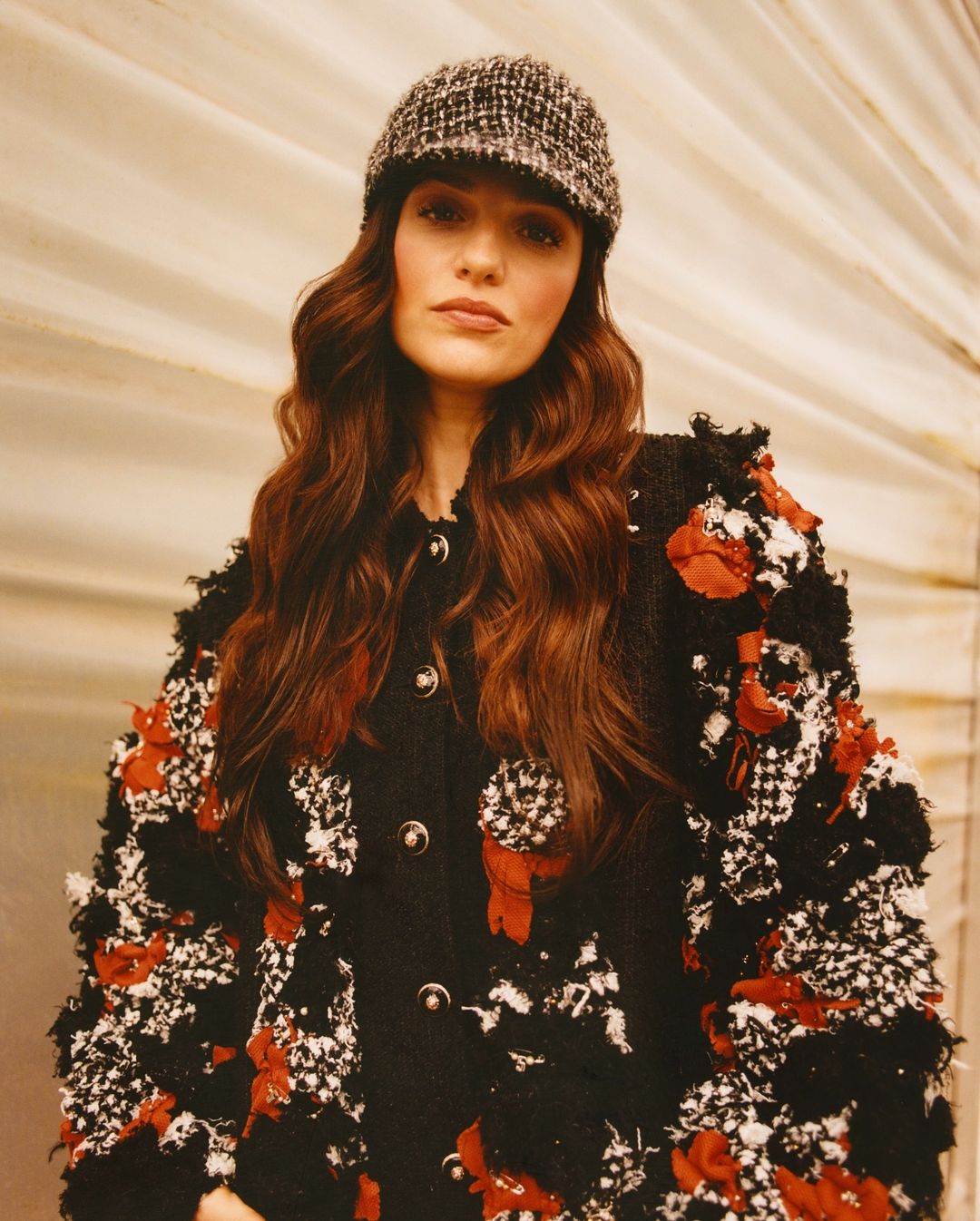 Elegant adult female wearing a textured black and white tweed jacket with red floral accents and a tweed cap, posing indoors in a studio.