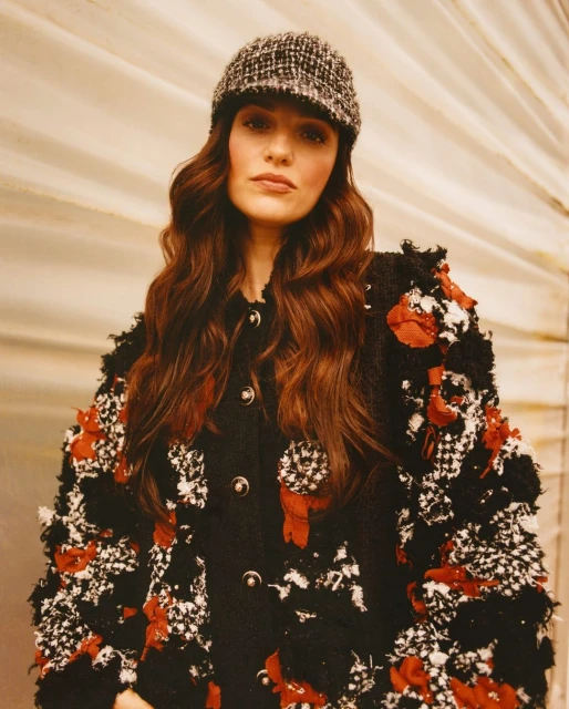 Elegant adult female wearing a textured black and white tweed jacket with red floral accents and a tweed cap, posing indoors in a studio.