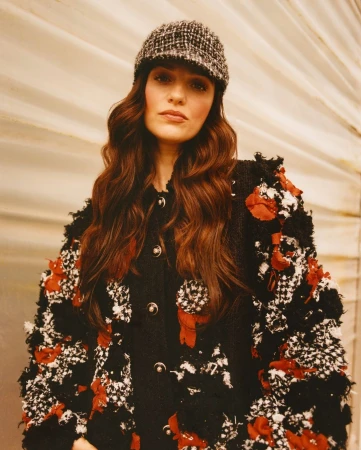 Elegant adult female wearing a textured black and white tweed jacket with red floral accents and a tweed cap, posing indoors in a studio.