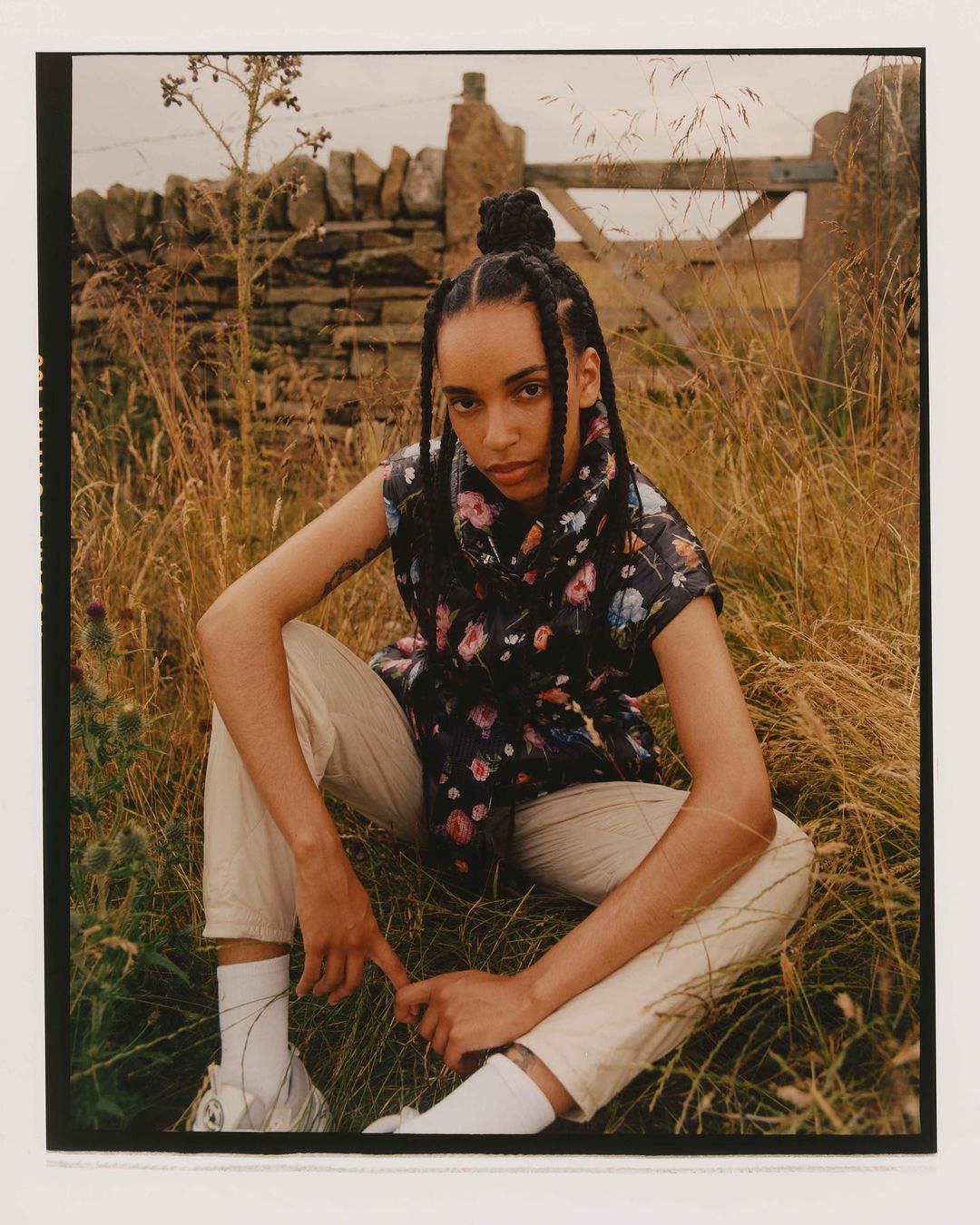 Woman in floral vest and light-colored pants sits in a grassy field with a stone wall and wooden gate in the background.