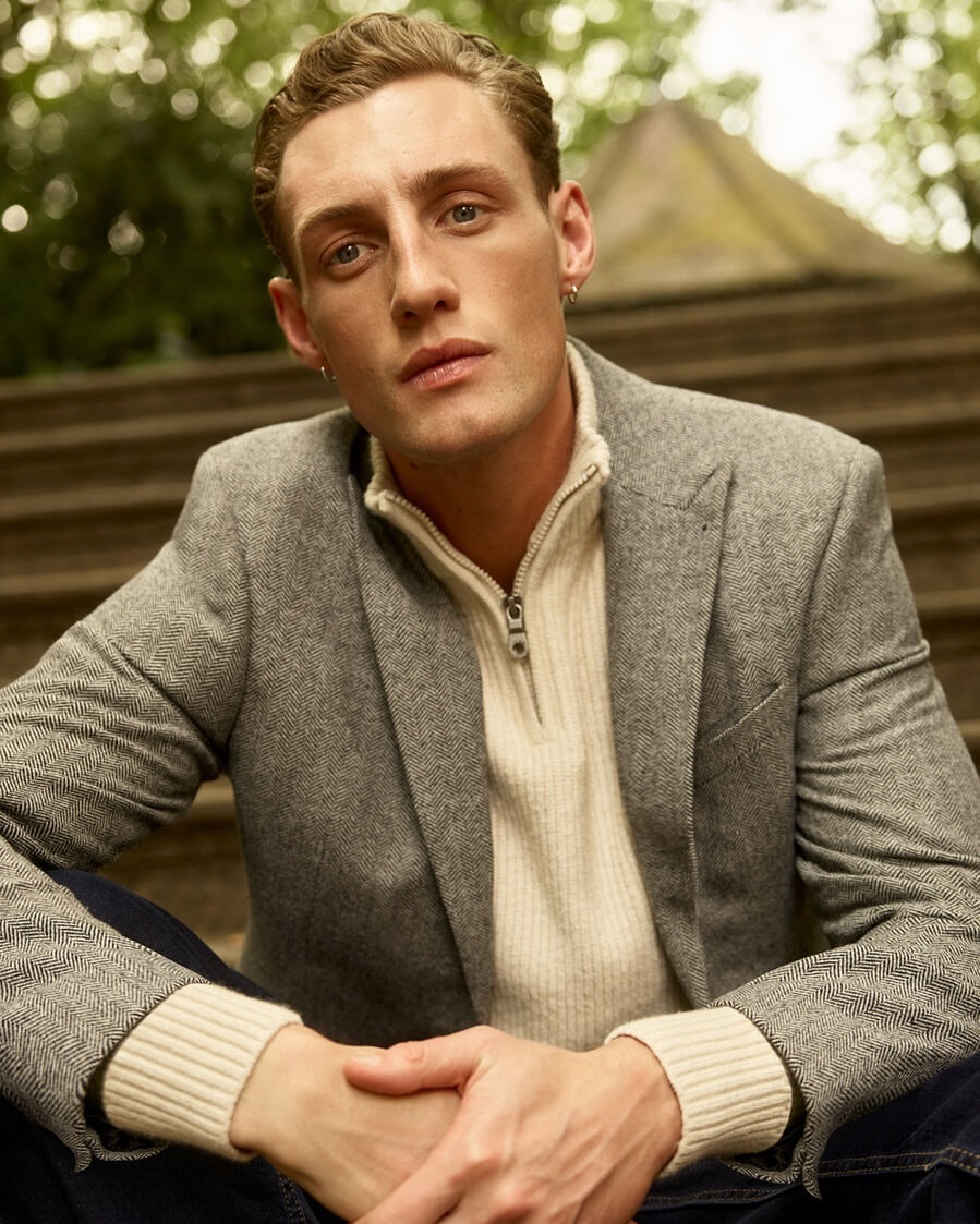 Portrait of a young adult male with blond hair wearing a blazer and collared shirt sitting on steps outdoors in soft, natural lighting.