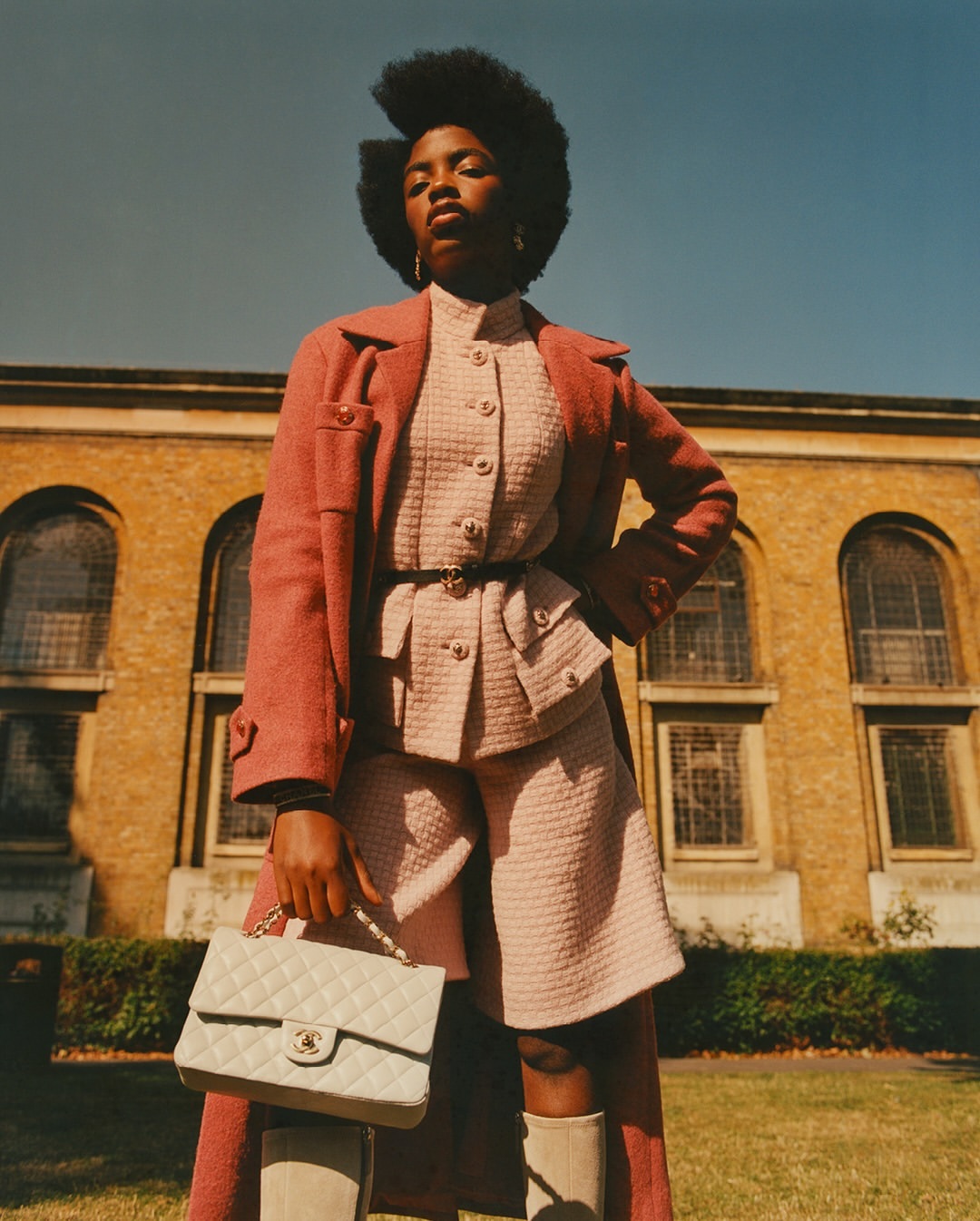 Fashionable Black woman poses in a pink tweed jacket and shorts set, accessorized with a cream handbag in front of a tan brick building.
