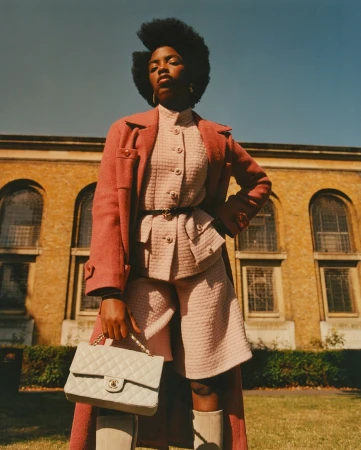 Fashionable Black woman poses in a pink tweed jacket and shorts set, accessorized with a cream handbag in front of a tan brick building.