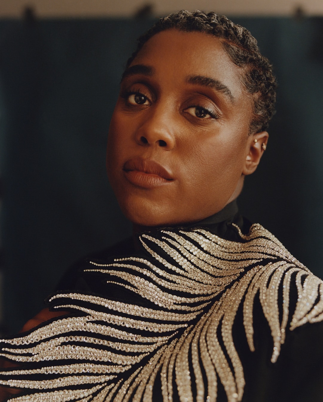Close-up portrait of a Black woman with short hair wearing a black top with silver beaded embroidery against a dark teal background.