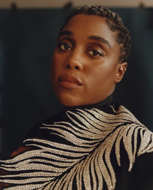 Close-up portrait of a Black woman with short hair wearing a black top with silver beaded embroidery against a dark teal background.