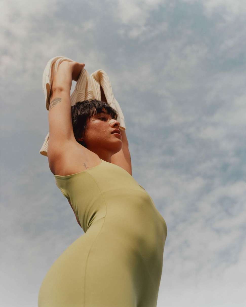 Woman in a light green dress stands outside against a blue sky, holding a cream cardigan above her head. Low angle shot.