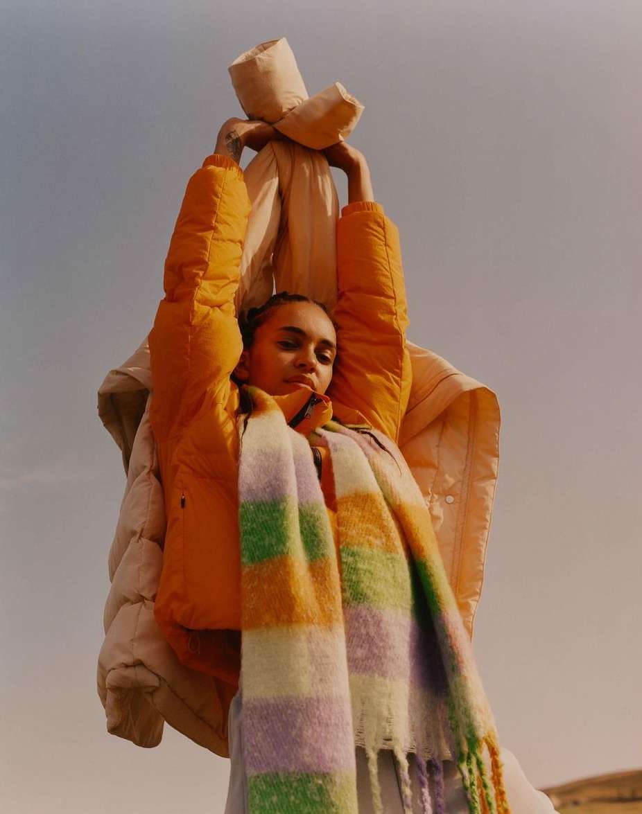 A young woman in a yellow puffer jacket poses outdoors against a sky background while holding a beige jacket overhead and wearing a colorful scarf.