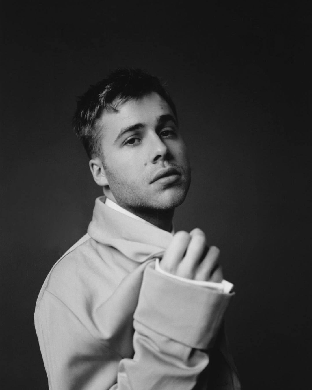 Monochrome portrait of a fair-skinned adult male looking at the camera and wearing a light-colored jacket against a dark grey backdrop.