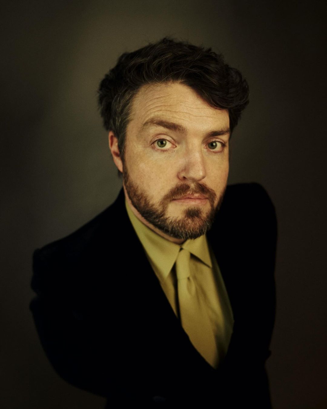 Portrait of a man with a beard wearing a dark blazer and tie against a dark background