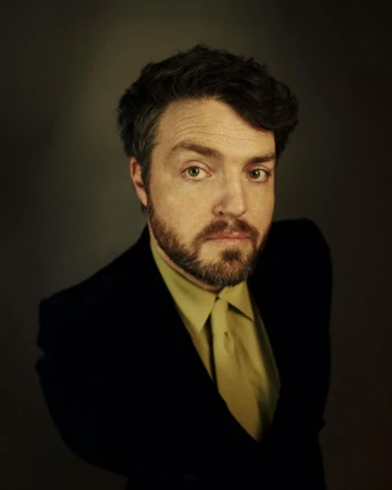 Portrait of a man with a beard wearing a dark blazer and tie against a dark background