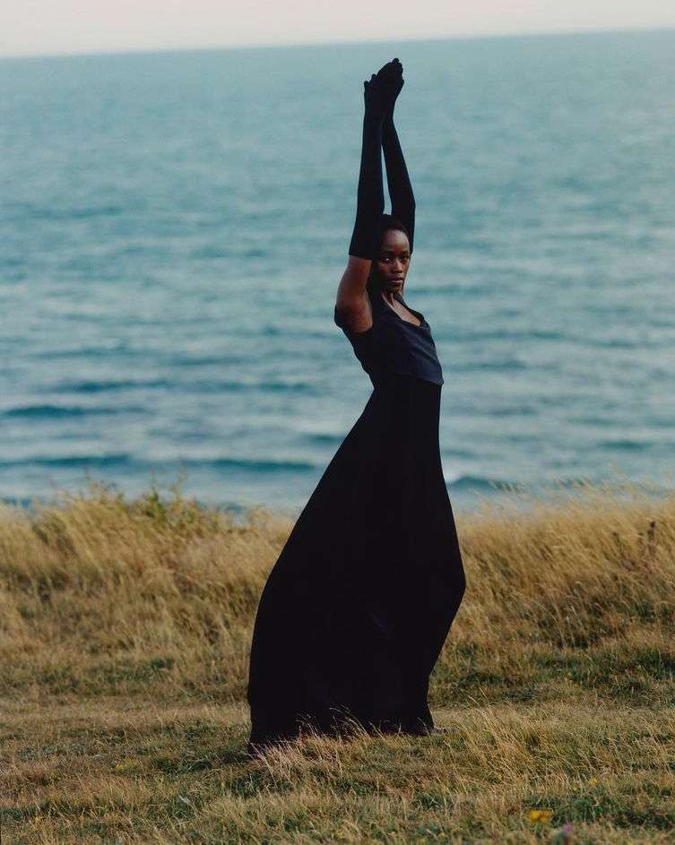 Woman in long black dress and gloves stands gracefully on a grassy hill overlooking the ocean, posing with arms raised overhead.