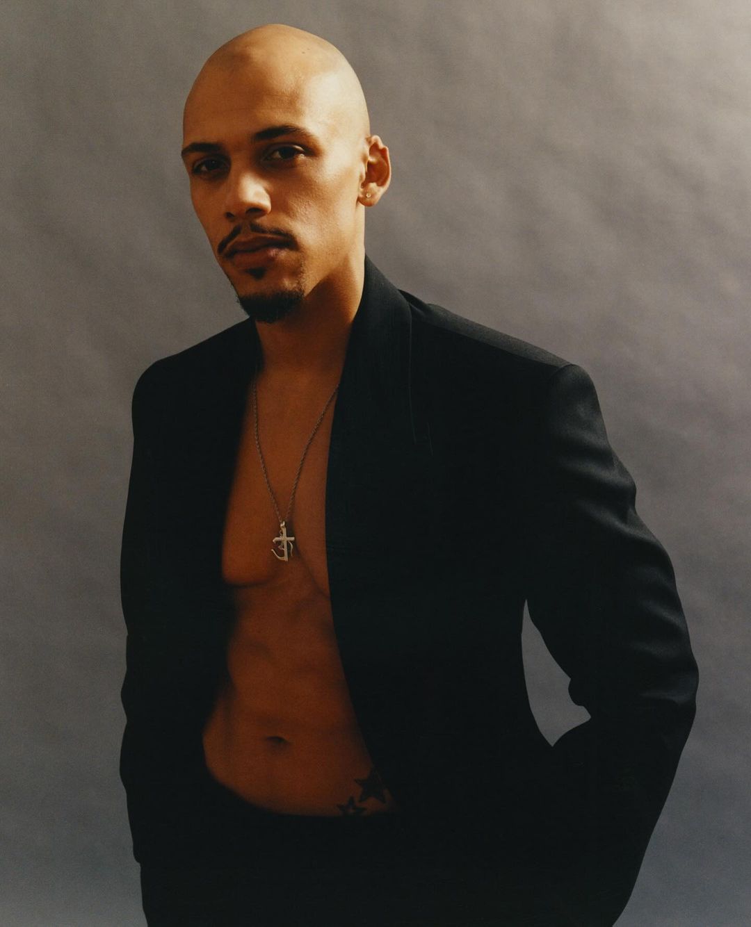 Portrait of a bald Black man wearing an open black blazer and a pendant necklace against a textured grey background.