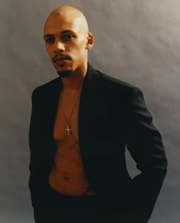 Portrait of a bald Black man wearing an open black blazer and a pendant necklace against a textured grey background.