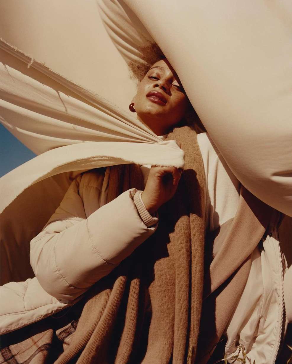 Woman poses outdoors wearing a beige jacket and scarf, partially covered by a flowing white sheet, capturing an artistic and ethereal feel.