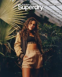 A woman with curly hair wearing a bomber jacket and high-waisted shorts posing in a lush tropical greenhouse setting.