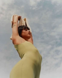 Low angle portrait of a woman in an olive green bodysuit holding a white knit garment up against a cloudy sky
