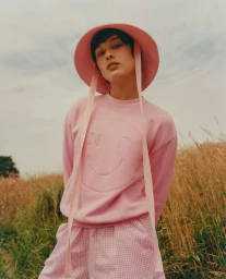 Model in pink sweatshirt and matching bucket hat standing in a tall grass field under an overcast sky.