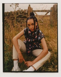 Woman in floral puffer vest and light trousers crouching in tall meadow grass with a stone wall behind her.