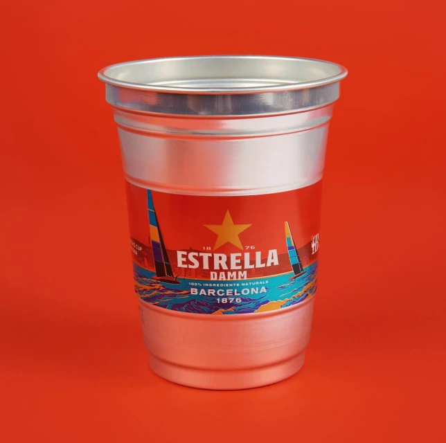 Still life photograph of an aluminum-colored cup featuring an Estrella Damm label against a solid orange background. Product shot.