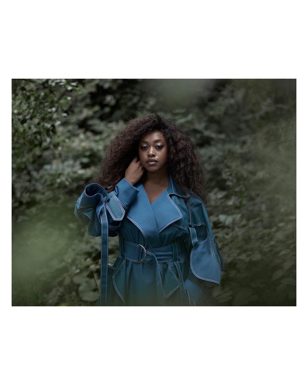 A woman with dark curly hair poses in a teal belted jacket outdoors, with a blurry green background creating a natural, soft setting.