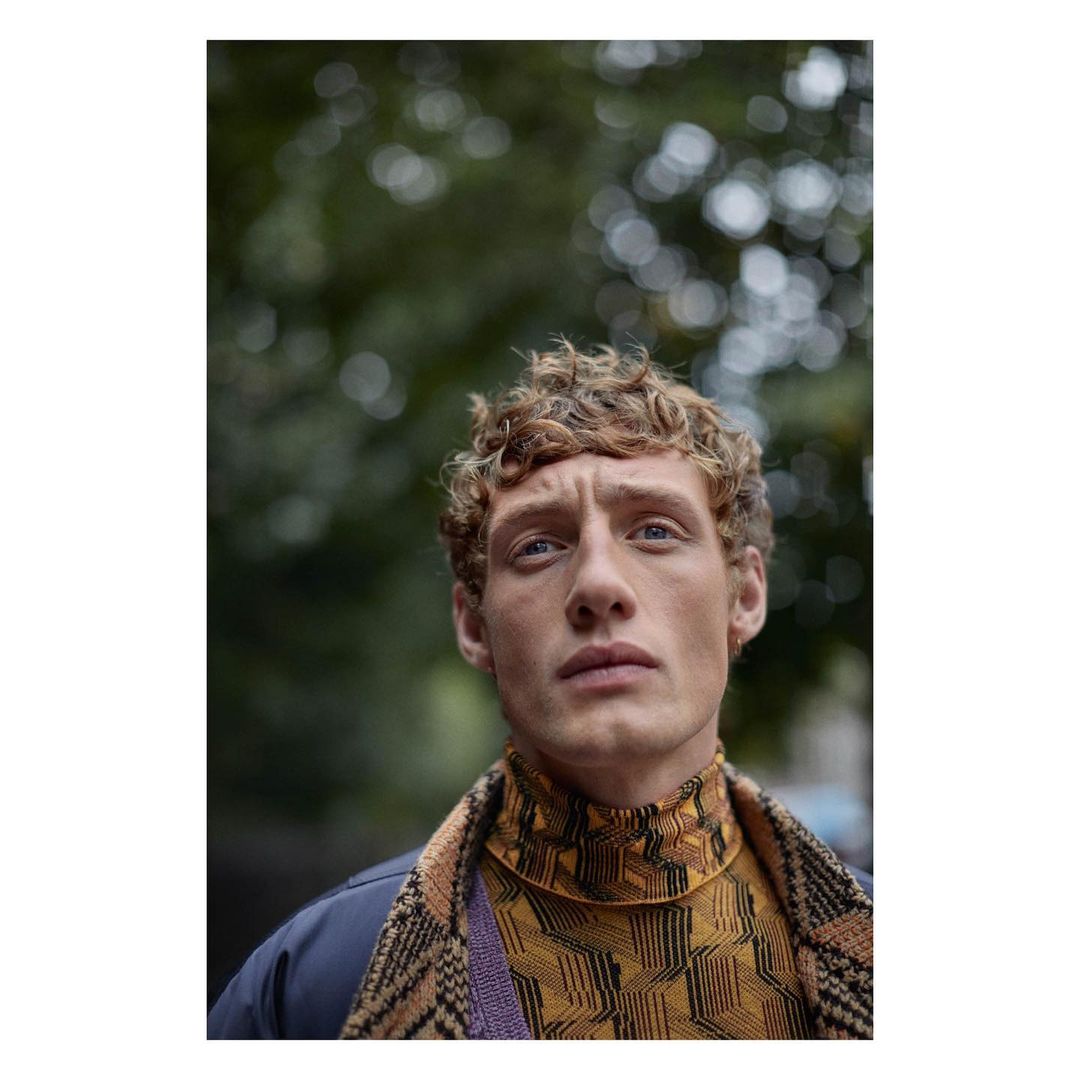 Portrait of a young adult male with curly ginger hair looking up, wearing a patterned turtleneck sweater and jacket against blurred green foliage.