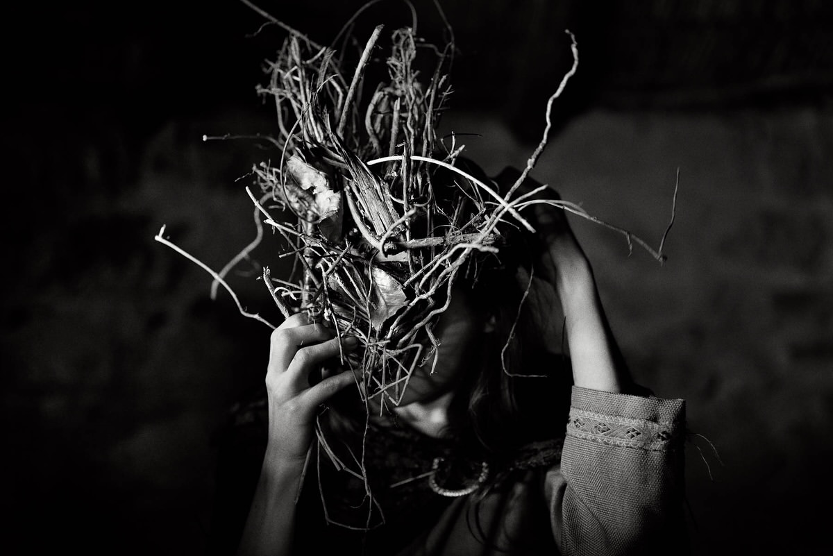 Monochrome portrait of a person wearing a large sculptural headdress made of tangled twigs and branches, photographed by Rich Gilligan.