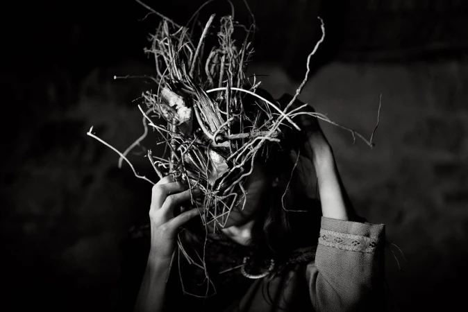Monochrome portrait of a person wearing a large sculptural headdress made of tangled twigs and branches, photographed by Rich Gilligan.