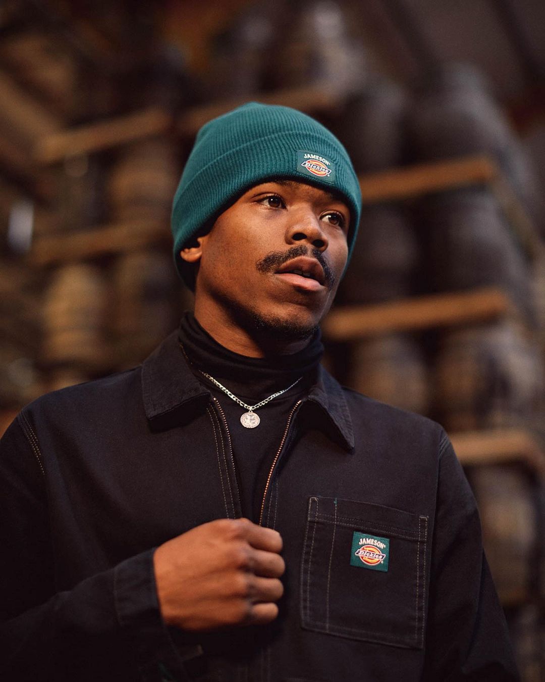 Portrait of a young adult man wearing a dark jacket and a teal beanie standing indoors with wood barrels in the background.