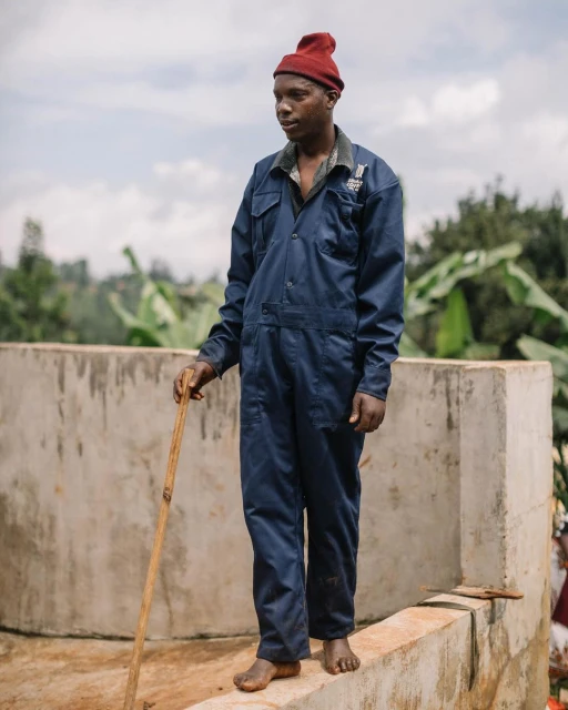 Barefoot man in blue work suit and red beanie holds a wooden stick while standing on a wall in a rural outdoor environment.