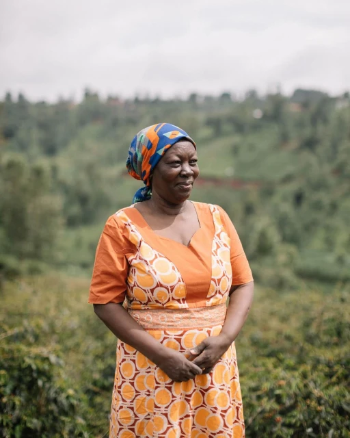 Portrait of a smiling African woman in a patterned orange dress and headscarf, standing in a green, hilly outdoor environment.