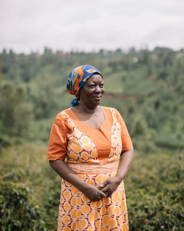 Portrait of a smiling African woman in a patterned orange dress and headscarf, standing in a green, hilly outdoor environment.