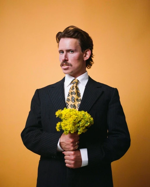 An adult man with a mustache, wearing a suit and tie, holds a bouquet of yellow flowers against an orange background.