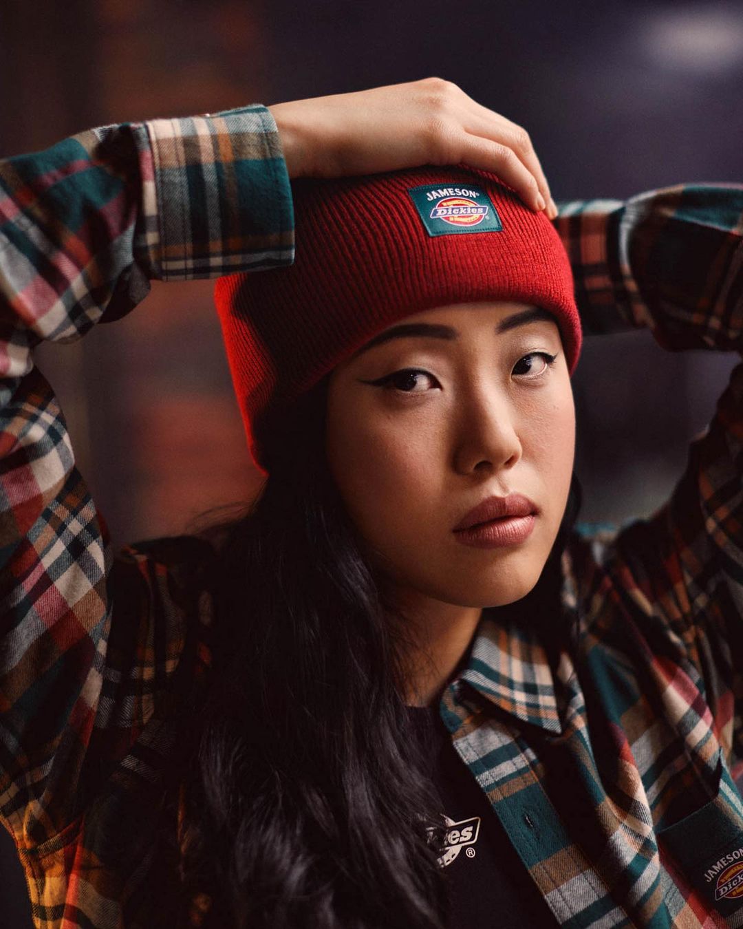 Portrait of a young Asian woman in a red beanie and plaid flannel shirt, hands on her head, gazing at the camera with a neutral expression.