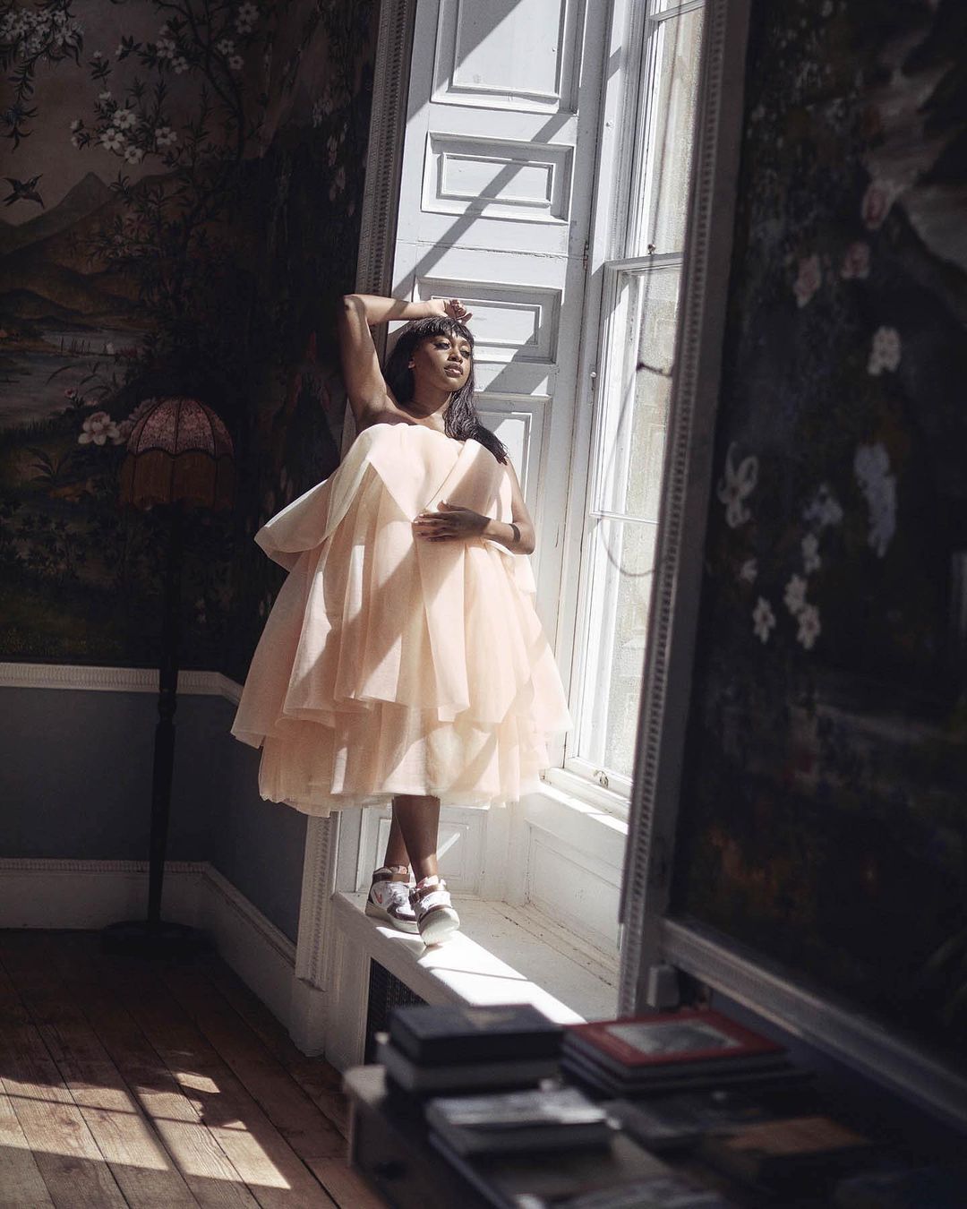 Woman in a ruffled dress poses near a window with floral wallpaper and stacked books in a brightly lit indoor setting.