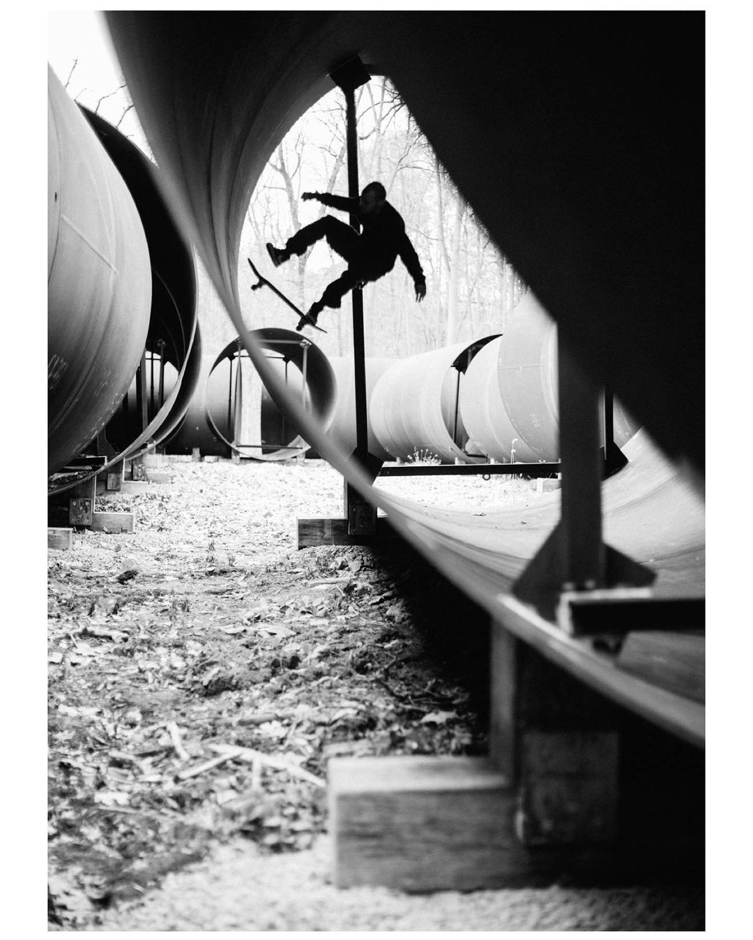 Monochrome shot of a skateboarder in mid-air performing a trick against a backdrop of pipes and trees. The image highlights action and silhouette.
