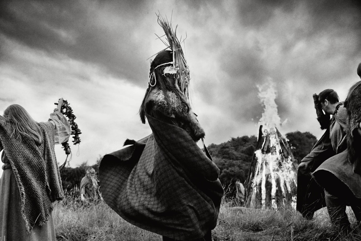 Monochrome photo of a group in ritualistic clothing around a large bonfire, set against a cloudy sky and grassy landscape.