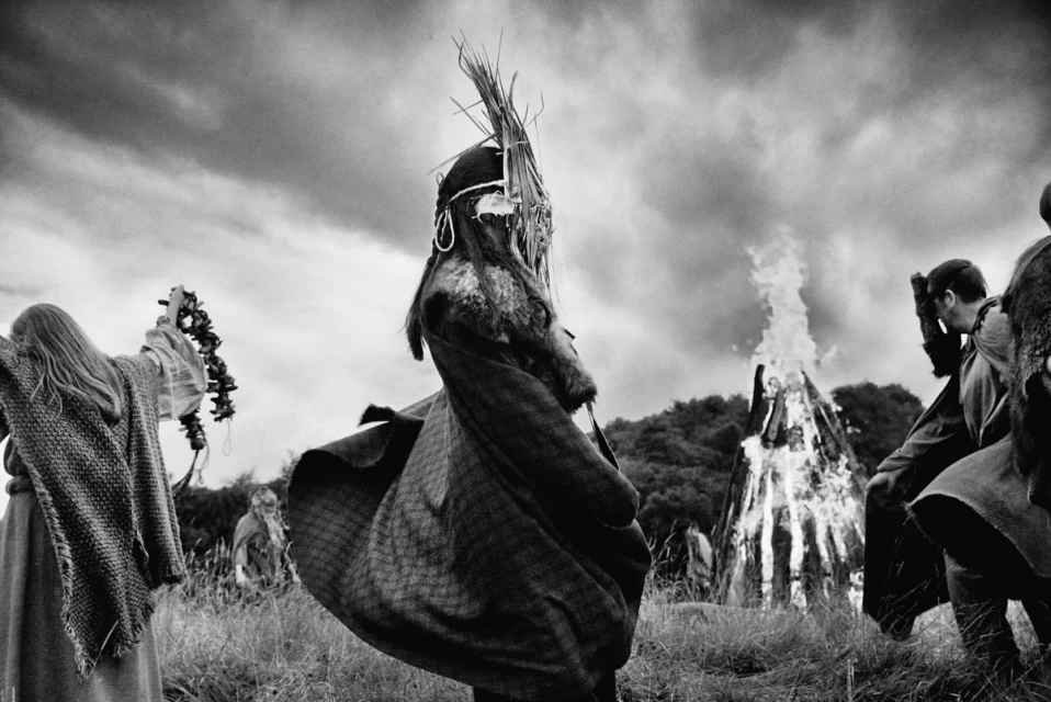 Monochrome photo of a group in ritualistic clothing around a large bonfire, set against a cloudy sky and grassy landscape.