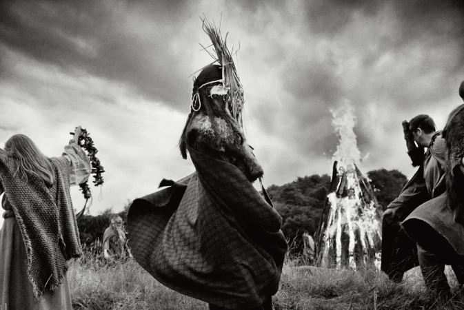 Monochrome photo of a group in ritualistic clothing around a large bonfire, set against a cloudy sky and grassy landscape.