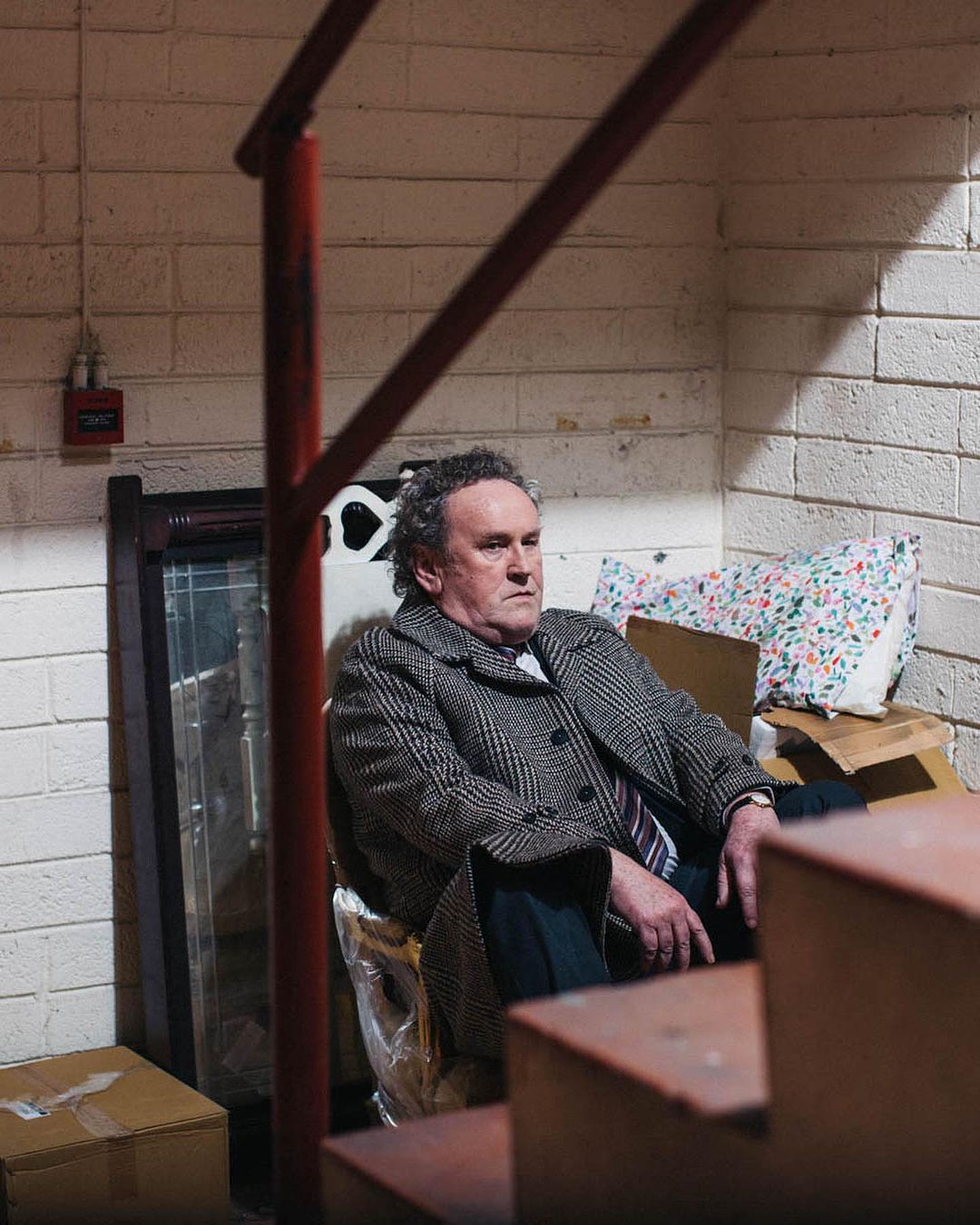 An adult male sits indoors on makeshift seating wearing a tweed overcoat and tie. A brick wall and red staircase railing are behind him.