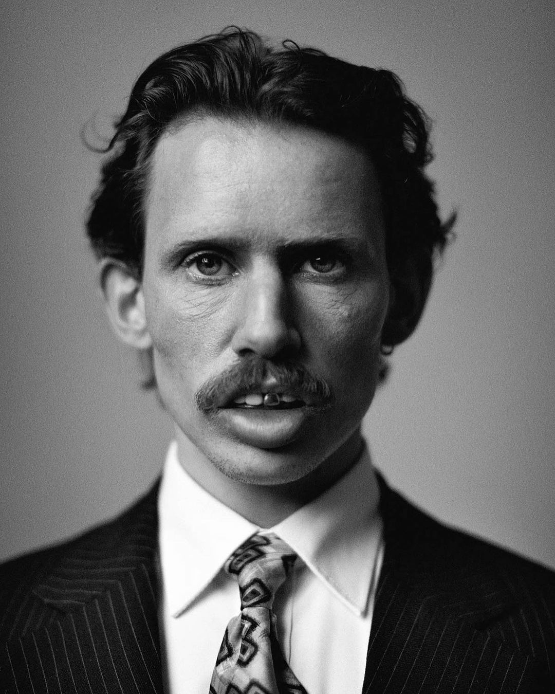 Black-and-white portrait of a man with a mustache wearing a pinstripe suit, white shirt, and patterned tie against a grey backdrop.