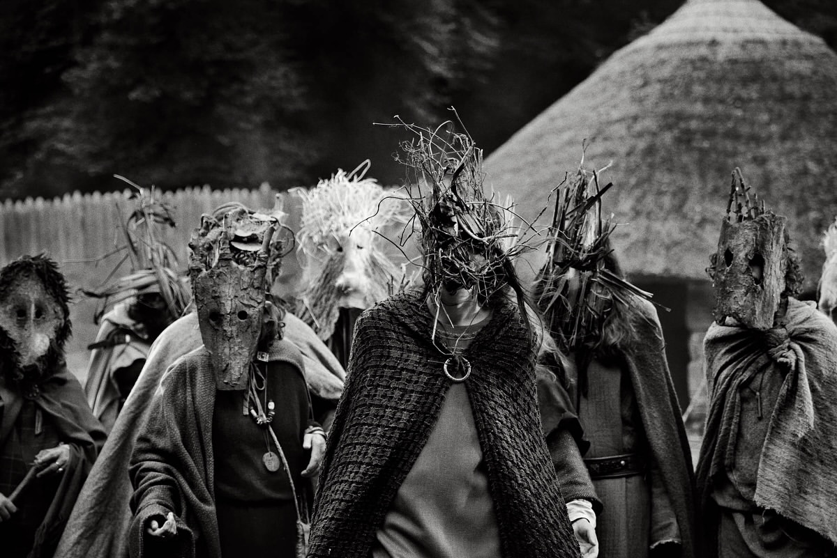 Monochrome image of a group of people wearing elaborate masks and draped in fabric, standing outdoors with a thatched-roof structure.