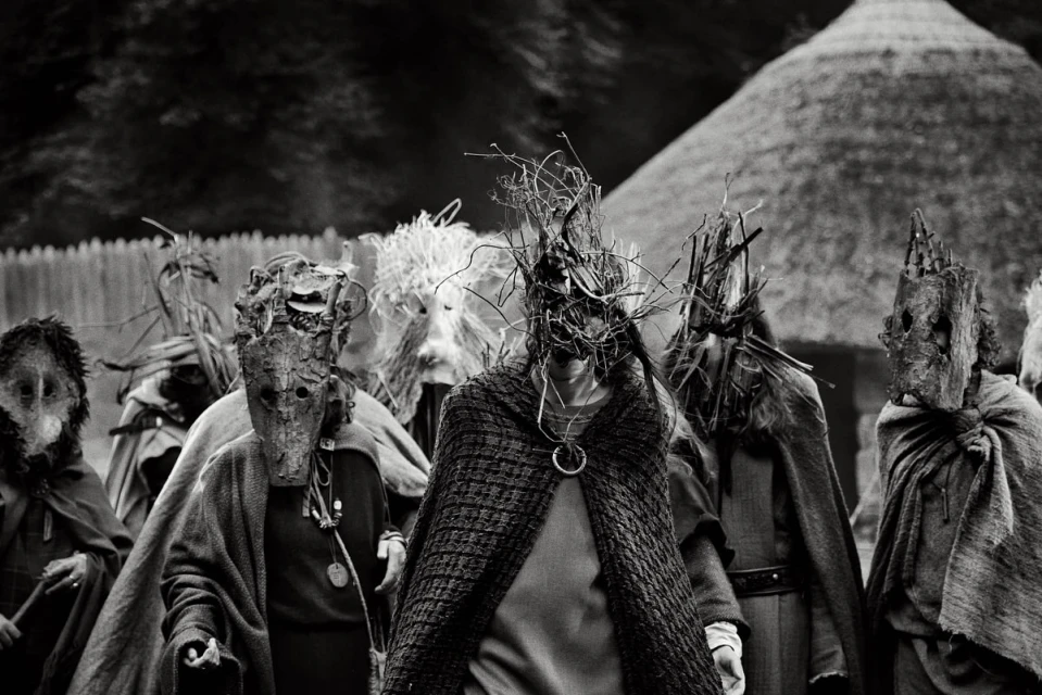 Monochrome image of a group of people wearing elaborate masks and draped in fabric, standing outdoors with a thatched-roof structure.