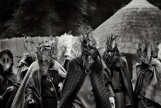Monochrome image of a group of people wearing elaborate masks and draped in fabric, standing outdoors with a thatched-roof structure.