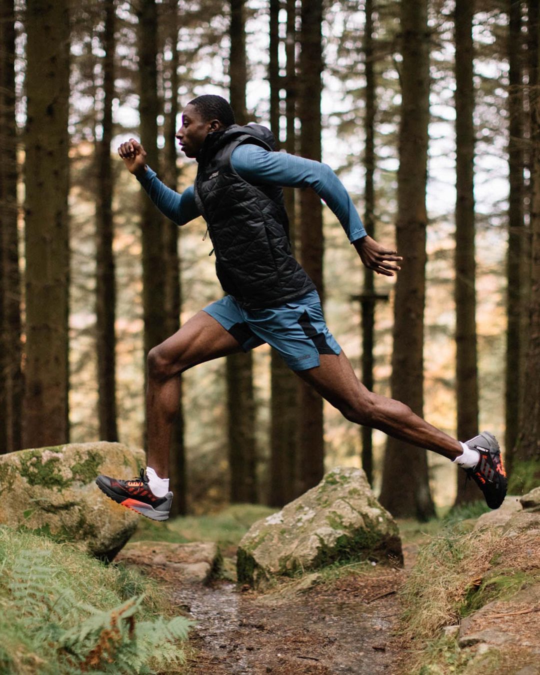Athletic Black man jumps over rocks during a trail run in a forest wearing a sleeveless puffer jacket and athletic shorts on a sunny day.