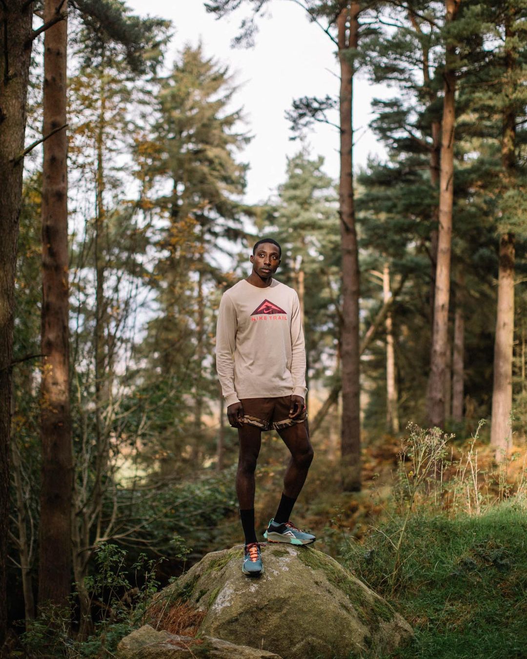 Athletic, dark-skinned man wearing a long sleeve shirt and shorts stands on a rock in a forest setting, looking at the camera.