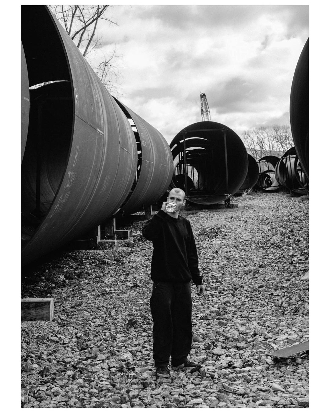 Monochrome portrait of an adult man in a black sweatshirt, holding a glass outside among large metal pipes and gravel under a cloudy sky.
