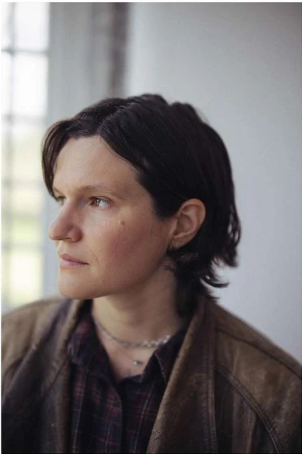 Portrait of a young adult female with short brown hair wearing a brown jacket and plaid shirt, posing indoors in front of a window.